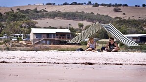 Een strand met een gestreepte parasol, een huis op de achtergrond en een glooiend landschap.