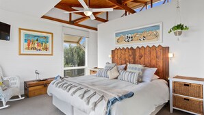 Een slaapkamer met een groot bed, een houten hoofdbord, een plafondventilator, een raam met uitzicht op bomen, en een aan de muur bevestigde televisie.