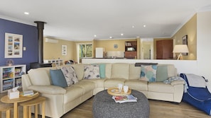 A living room with a beige sectional sofa, a round ottoman, and a coffee table. There is a bookshelf with books and games, a kitchen area with a refrigerator and a clock, and a dining area with a table and chairs.