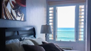 Une chambre à coucher avec un lit, des oreillers, un tableau, une fenêtre avec des volets et une vue sur la mer.