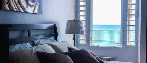 Ein Schlafzimmer mit Bett, Kissen, einem Gemälde, einem Fenster mit Rollläden und Blick aufs Meer.