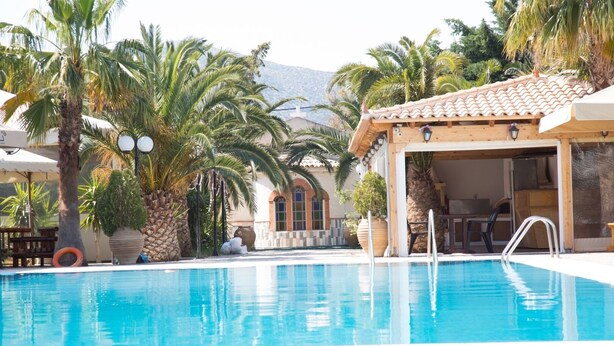 A pool area with a covered seating area, palm trees, and a mountain in the background. - One of the biggest Villas Near Athens Greece 40% off January 1 to May 15 (ANAVISSOS)