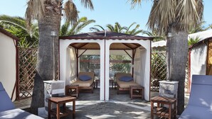 A covered outdoor seating area with two lounge chairs and a table, surrounded by palm trees and a stone floor. - One of the biggest Villas Near Athens Greece 40% off January 1 to May 15 (ANAVISSOS)