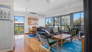 A spacious living area with a grey sofa, wooden coffee table, and a dining table with chairs. Large windows provide ample natural light and views of the outdoors.