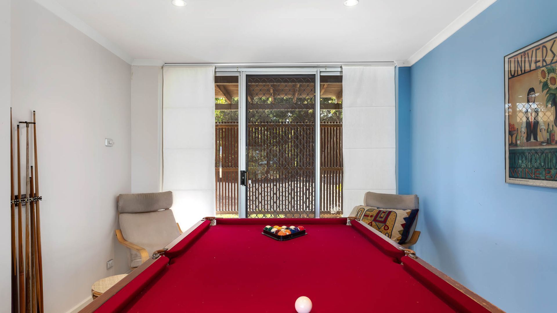 A pool table with a red felt surface, two chairs, and a window with a view of greenery.