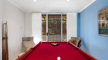 A pool table with a red felt surface, two chairs, and a window with a view of greenery.