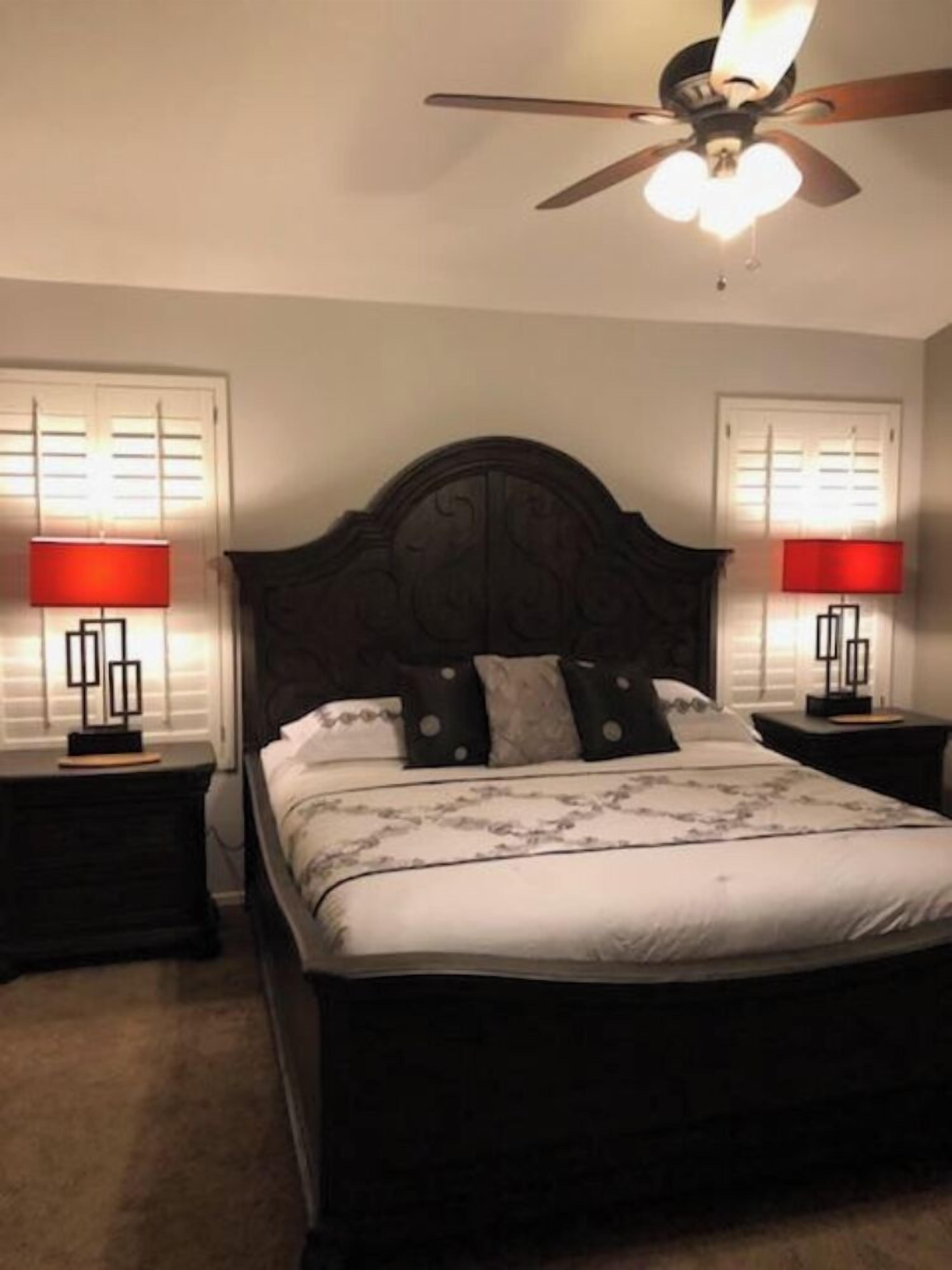 A bedroom with a large bed, two bedside tables with lamps, a ceiling fan, and windows with blinds.