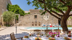 Area pranzo all'aperto con muri in pietra, piscina e un albero che offre ombra.