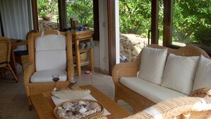 Un espace extérieur accueillant, aménagé avec des meubles en osier, une table ornée d’un bol rempli de coquillages, et offrant une vue sur une végétation luxuriante grâce à de grandes fenêtres.