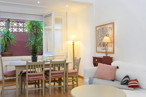 A cozy living room with a dining area, a sofa with patterned pillows, a lamp, and a window with plants.
