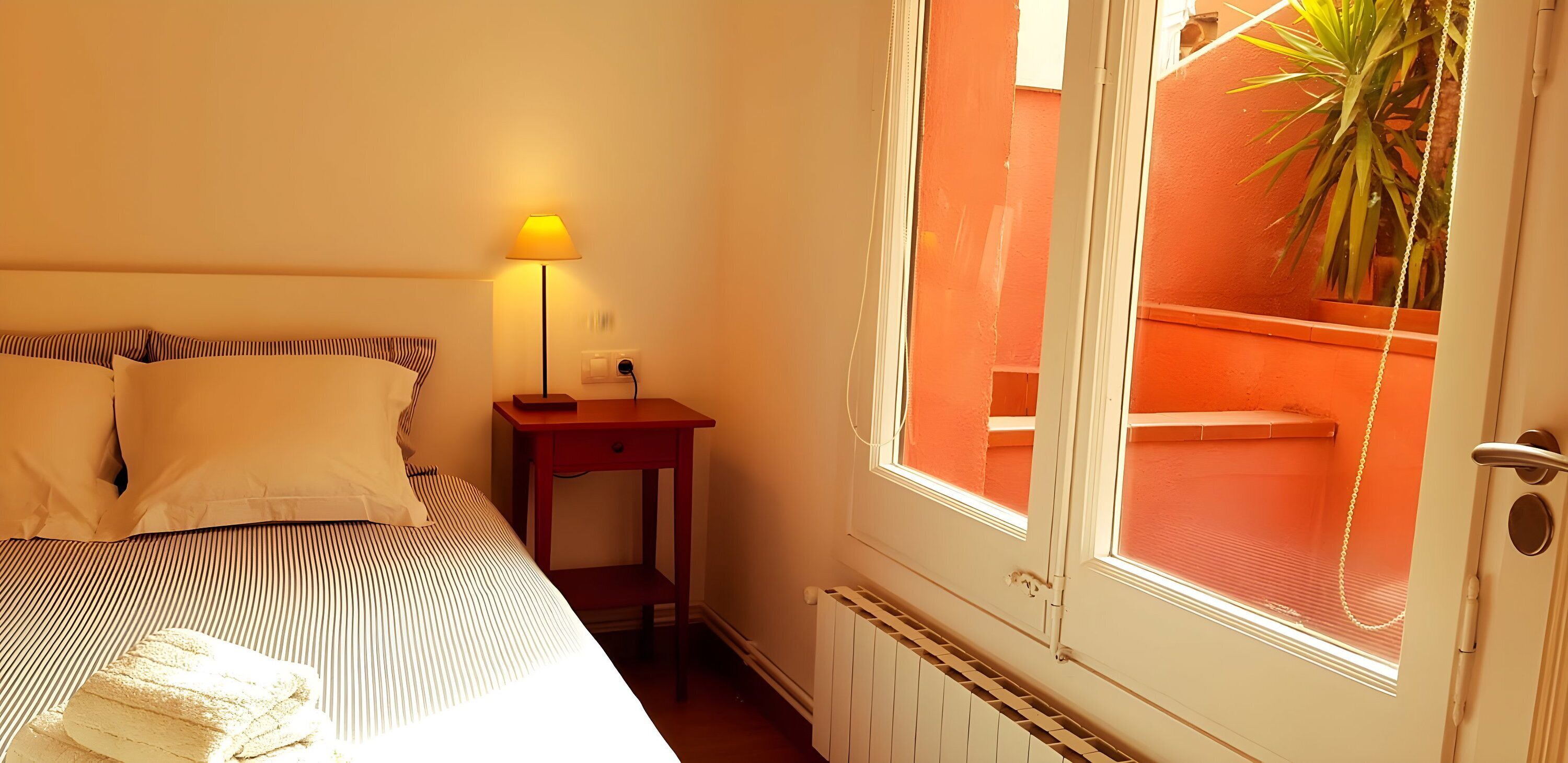 Et soveværelse med en seng, et natbord med lampe, en radiator og et vindue med udsigt til en orange væg og en plante.