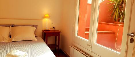 Et soveværelse med en seng, et natbord med lampe, en radiator og et vindue med udsigt til en orange væg og en plante.
