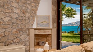 Un muro de piedra, una chimenea con un cuadro encima, una mesa de madera con una botella y un cuenco, y vistas al mar y las montañas a través de un umbral abierto.