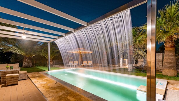 Una piscina al aire libre con una cascada, rodeada de sillones de descanso y un porche cubierto.