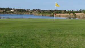 Um campo de golfe com haste de sinalização em primeiro plano, um corpo de água e uma paisagem acidentada ao fundo.