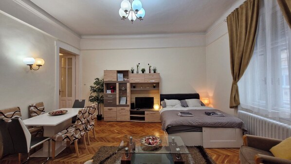 A compact hotel room with a bed, a desk, a chair, a TV, and a small dining table. - River Center Apartment (XIII. kerület)