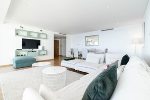 Una sala de estar moderna con un sofá blanco, un reposapiés azul, una mesa de centro blanca y un televisor sobre una estantería verde.