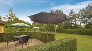 An outdoor patio with a table and chairs, a large umbrella, and neatly trimmed hedges.