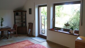 A room with a dining table, chairs, a bookshelf, a window with a view of greenery, and a small shelf with decorative items. - Hamburg Rahlstedt / Farmsen, small but very fine (Wandsbek)