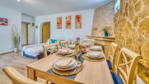 Un coin repas avec une table en bois, dressée pour quatre personnes, entourée de chaises. Un mur en pierre se trouve d’un côté, et un lit avec une tête de lit blanche est visible au fond.