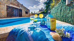 Une scène de bord de piscine avec une table où l’on aperçoit des boissons, une bouteille de gin Bombay Sapphire et une canette de Schweppes.
