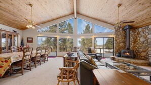 A spacious living area with a high ceiling, large windows, a stone fireplace, and a dining area with wooden furniture.