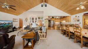 A spacious living area with a high ceiling, wooden beams, a dining table with chairs, a sofa, a television, and a kitchen in the background.