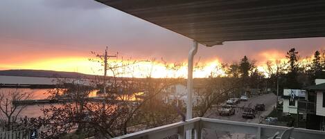 A balcony with a view of a sunset over a body of water, a street with parked cars, and a sign that reads "Bayfield".