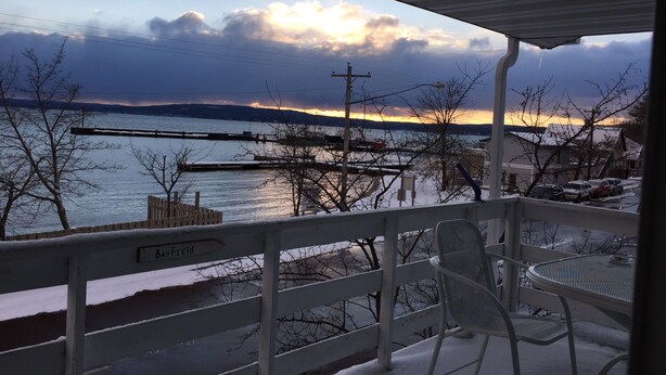 A covered outdoor area with a view of a body of water and a dock. There is a chair and a table, and a sign that reads "Bayfield". - Escape To Madeline Island !  Superior Lakefront 2 Bedroom Condo  (La Pointe)