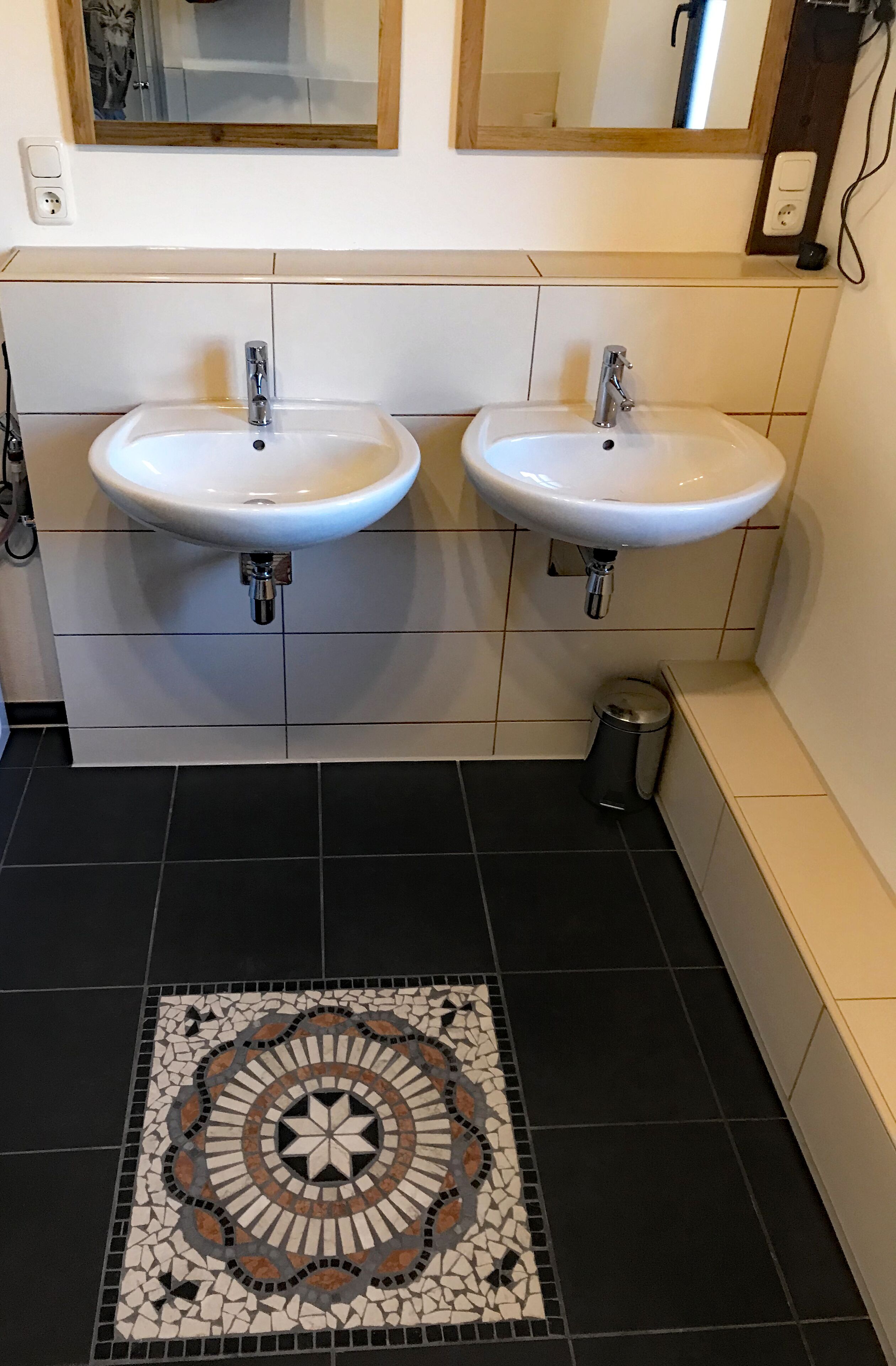 Ein Badezimmer mit zwei weißen Waschbecken, einem gefliesten Boden mit Dekor und einer kleinen Mülltonne.