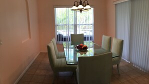 A dining area with a glass table surrounded by four chairs, a fruit bowl on the table, and a window with blinds. - Charmer in the Heart of Cape Coral (Cape Coral)
