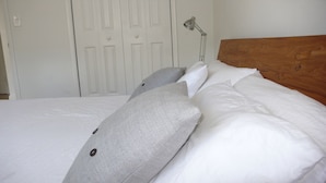 Een opgemaakt bed met witte lakens en grijze kussens, een houten hoofdbord en een witte deur op de achtergrond.
