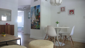 Een moderne eethoek met een witte tafel, stoelen en een potplant. Aan de muur hangt een kleurrijk schilderij, en er staat een houten kast met een spiegel erboven.