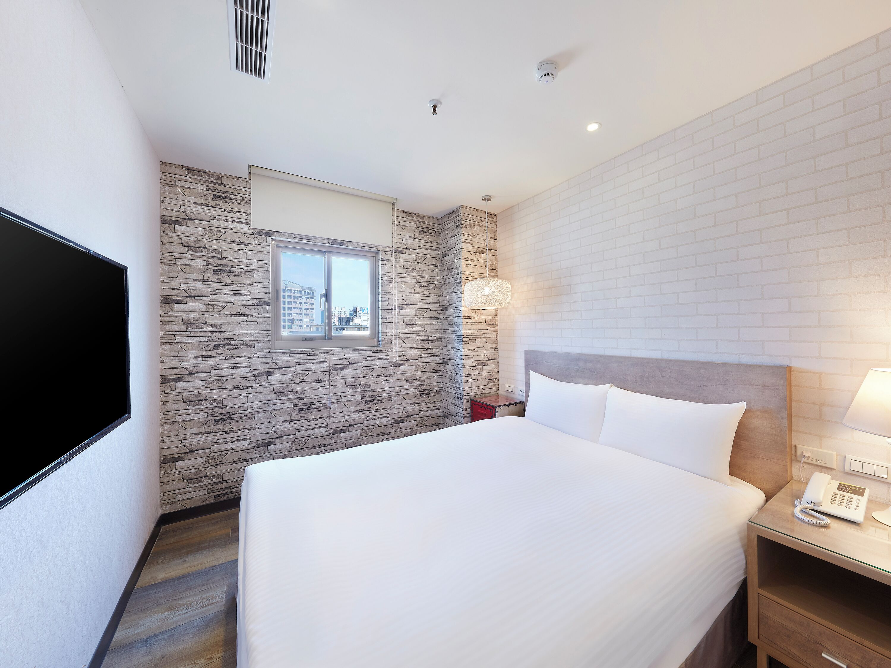 A hotel room with a large bed, a flat-screen TV, a stone wall, and a window with a city view.