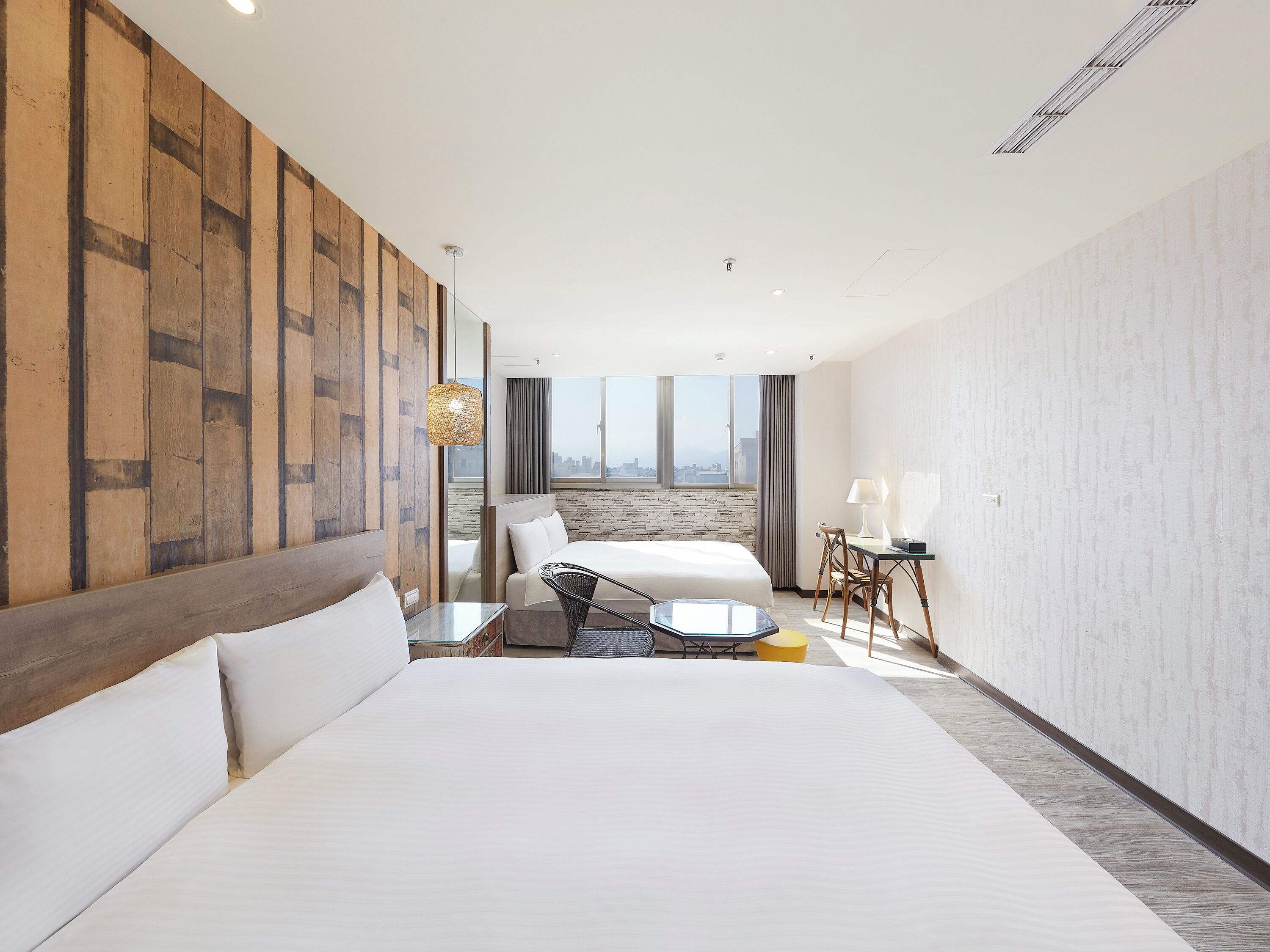 A hotel room with a large bed, a desk with a chair, a view of the city through a large window, and a wooden accent wall.