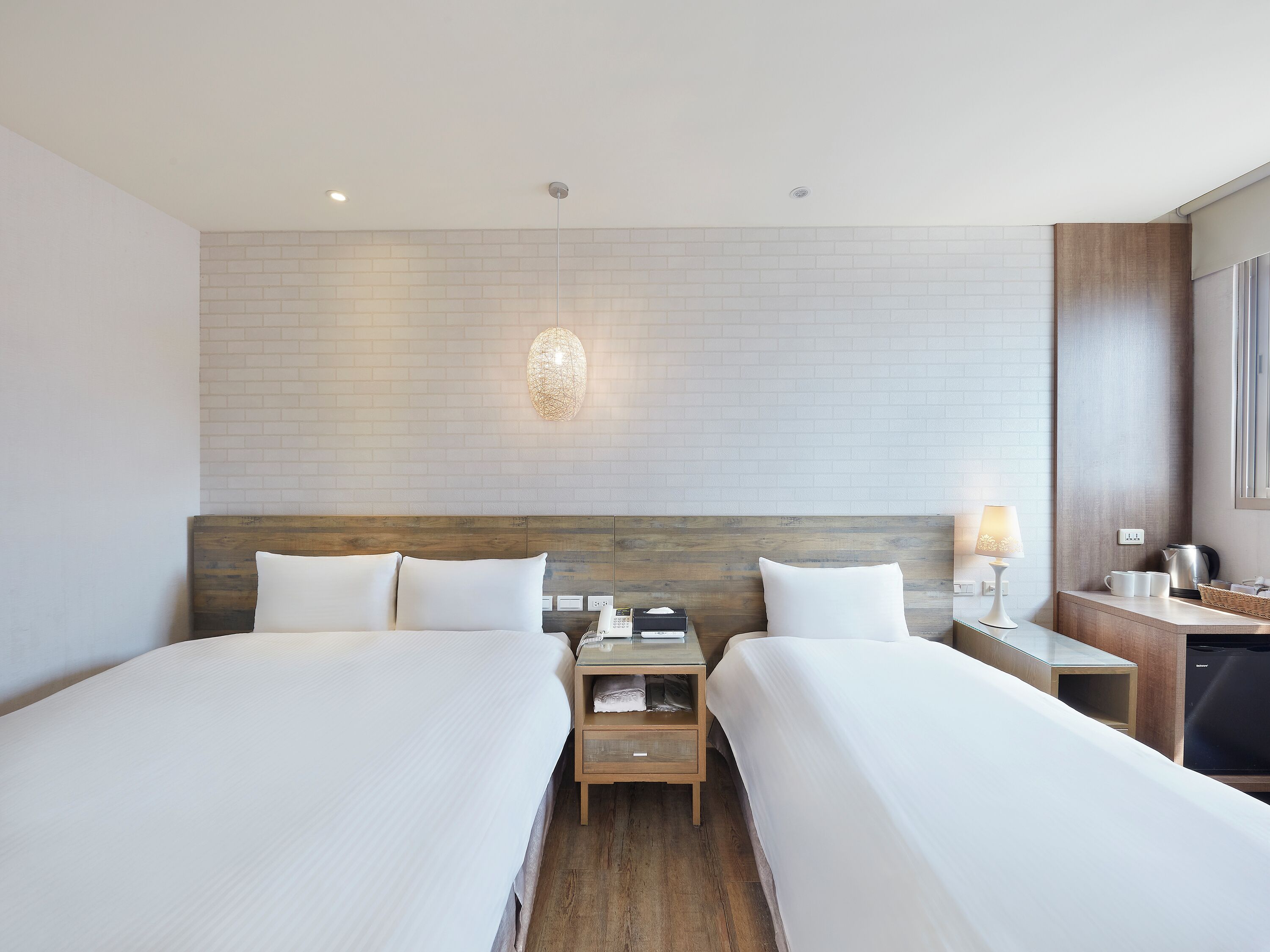 Habitación de hotel con dos camas, cabecera de madera, mesita de noche con lámpara, pequeña mesa con hervidor y pared con diseño de ladrillos.