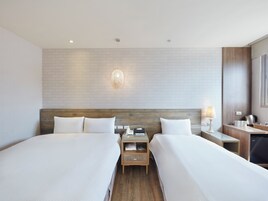 Habitación de hotel con dos camas, cabecera de madera, mesita de noche con lámpara, pequeña mesa con hervidor y pared con diseño de ladrillos.