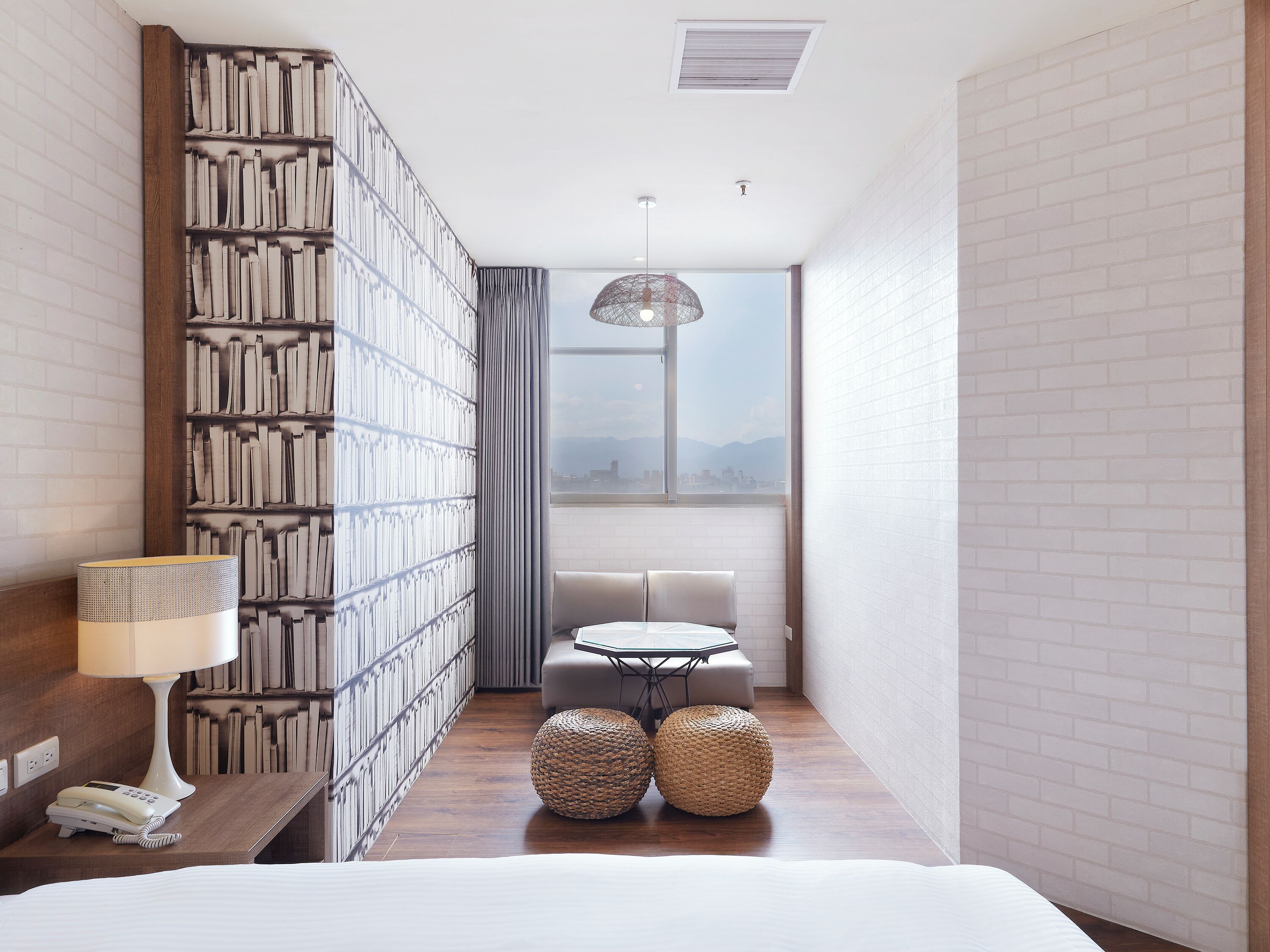 A hotel room with a bed, a desk with a lamp, a bookshelf, a round woven ottoman, and a view of the city through a large window.