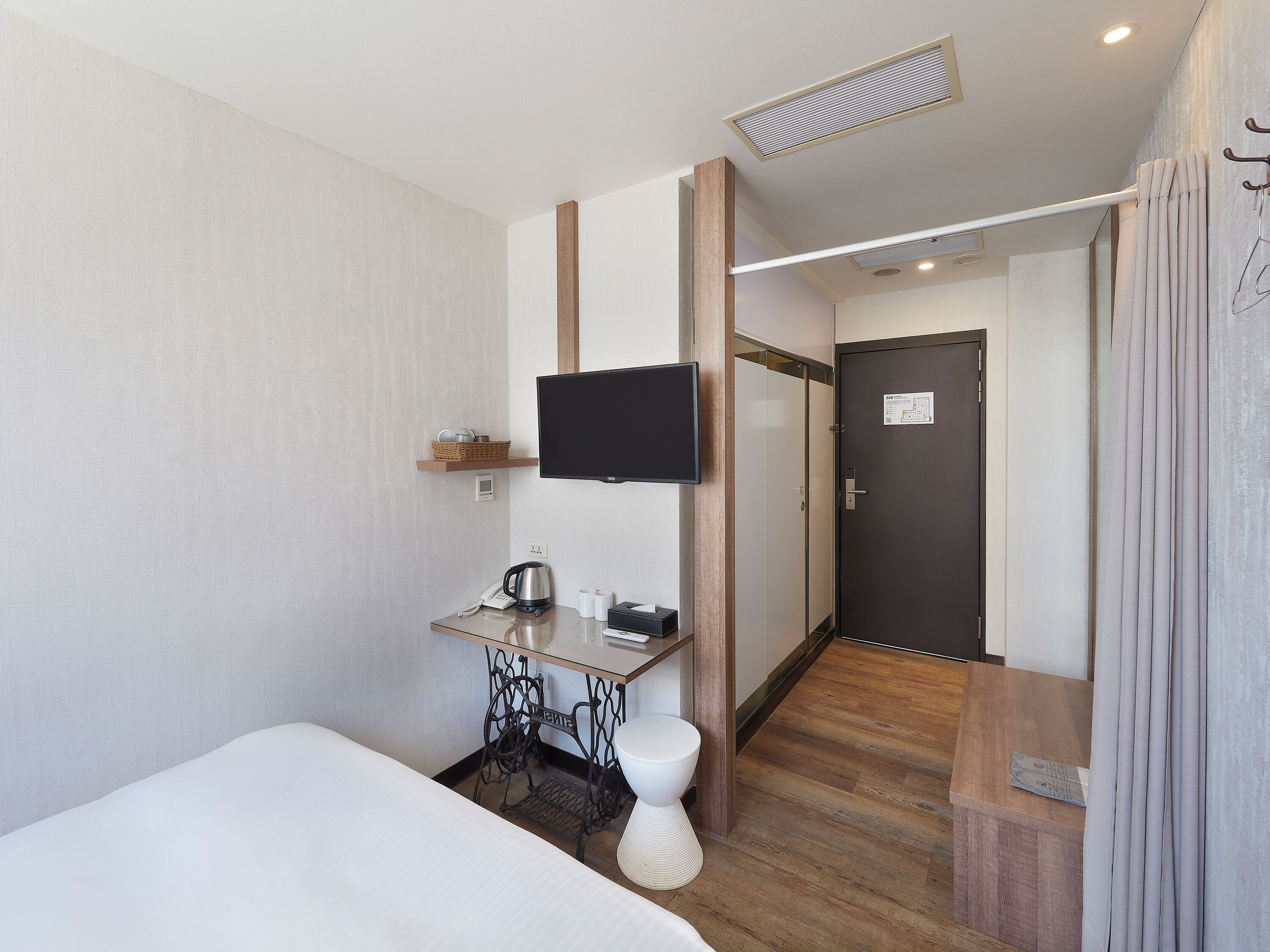 A hotel room with a bed, a desk with a telephone and a kettle, a television mounted on the wall, and a door with a sign.