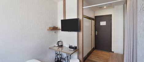 A hotel room with a bed, a desk with a telephone and a kettle, a television mounted on the wall, and a door with a sign.