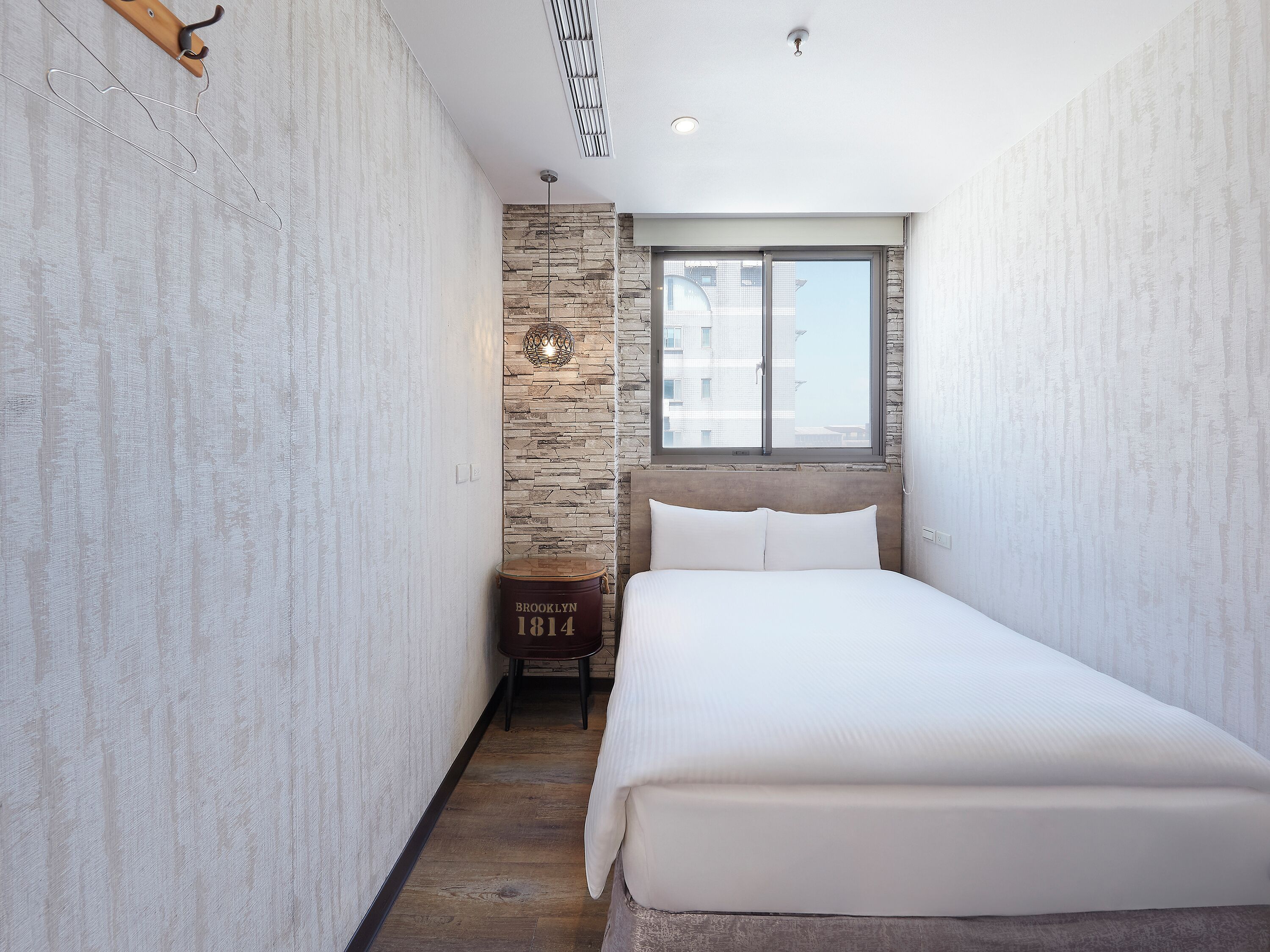Una habitación de hotel estrecha con una cama, un muro decorativo de piedra, una ventana y una lámpara fijada a la pared.