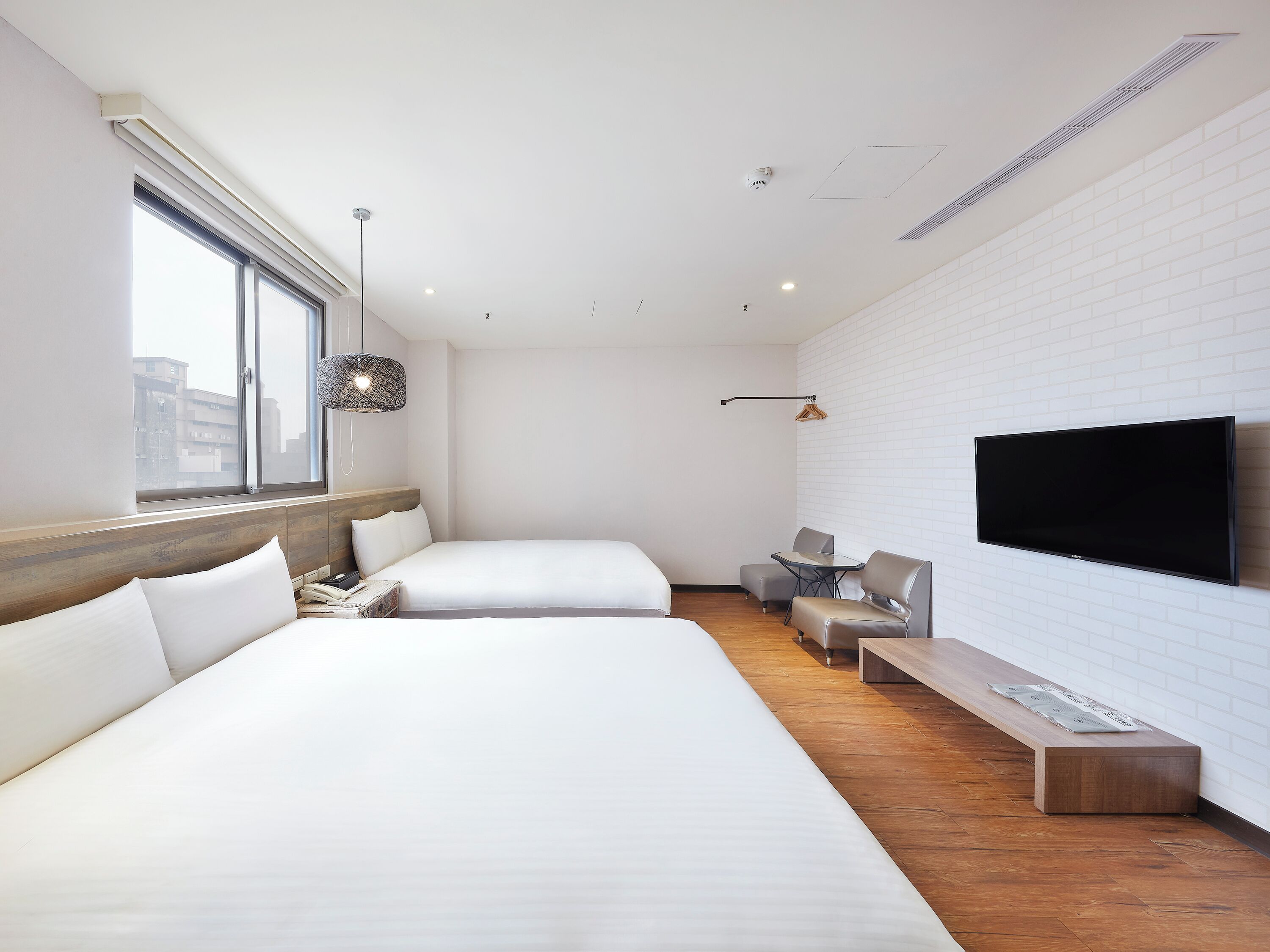 A modern hotel room with two beds, a flat-screen TV, a small table, and a window with a view of buildings.