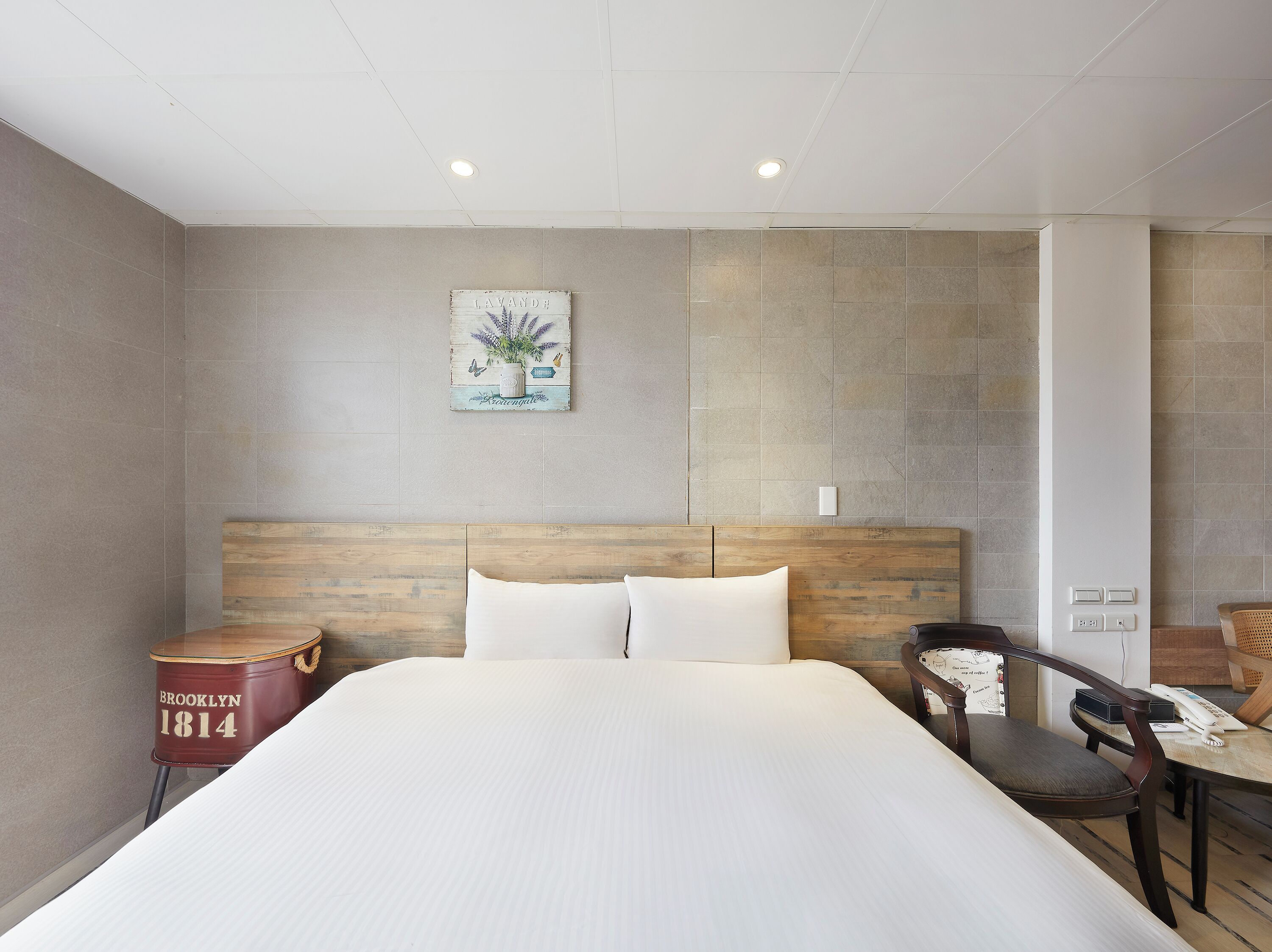 A hotel room with a large bed, a wooden headboard, a small table with a chair, and a wall-mounted artwork.