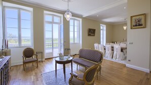A spacious room with a large window, a chandelier, a dining area with chairs, and a sofa.