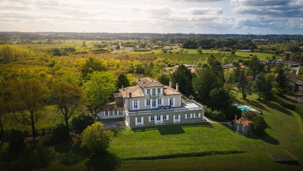A large, two-story house with a tiled roof, surrounded by lush green fields and trees.