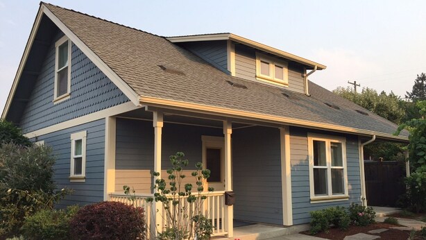 A two-story house with a gabled roof, a front porch with a railing, and a small garden with various plants and flowers. - BLOSSOM VIEW COTTAGES: Luxurious 3 Bedroom + Den  Near Theaters, Downtown (Ashland)