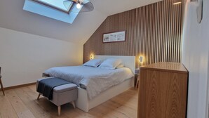 Quarto moderno com teto inclinado, cama com colcha estampada em padrão geométrico, um pequeno banco, mesa de cabeceira de madeira e luminária fixada na parede.
