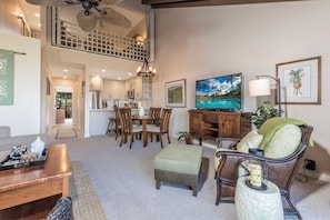 A spacious living area with a dining table, comfortable seating, a television, and a view of the kitchen and upper level.