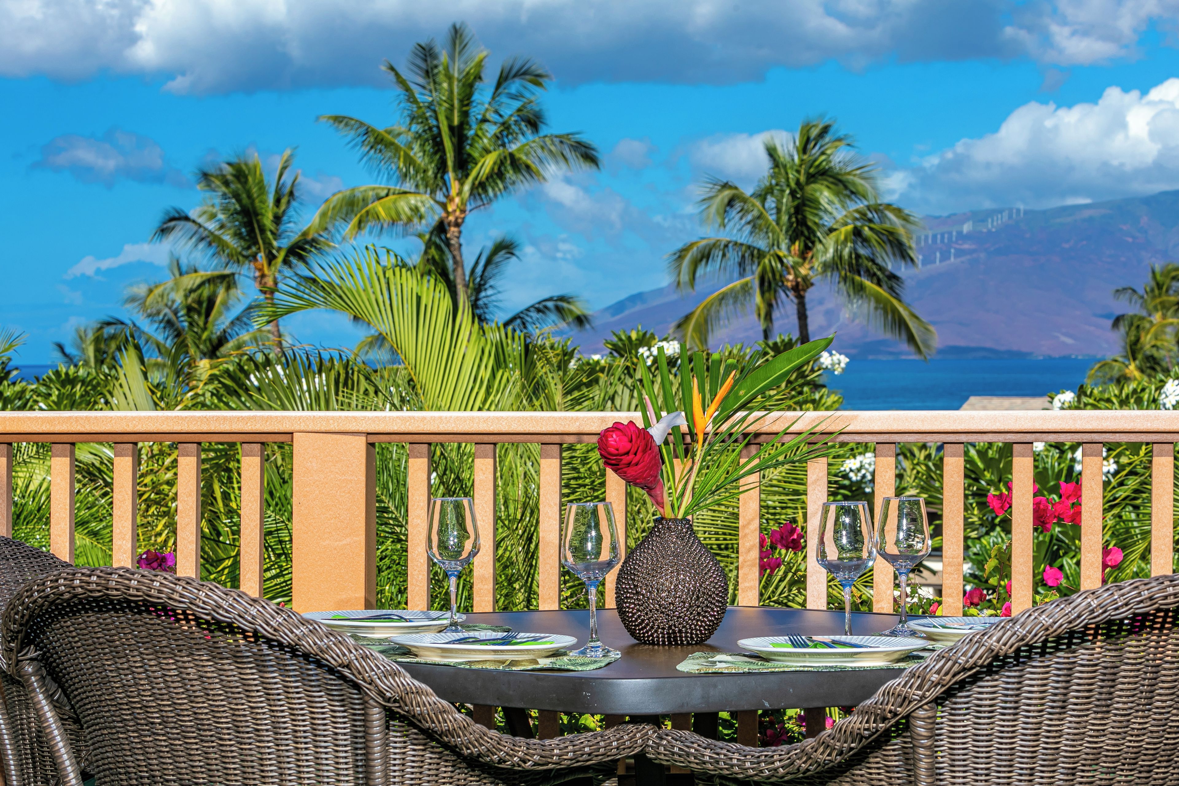 A dining table set with wine glasses and a vase, overlooking a tropical landscape with palm trees and a view of the ocean.