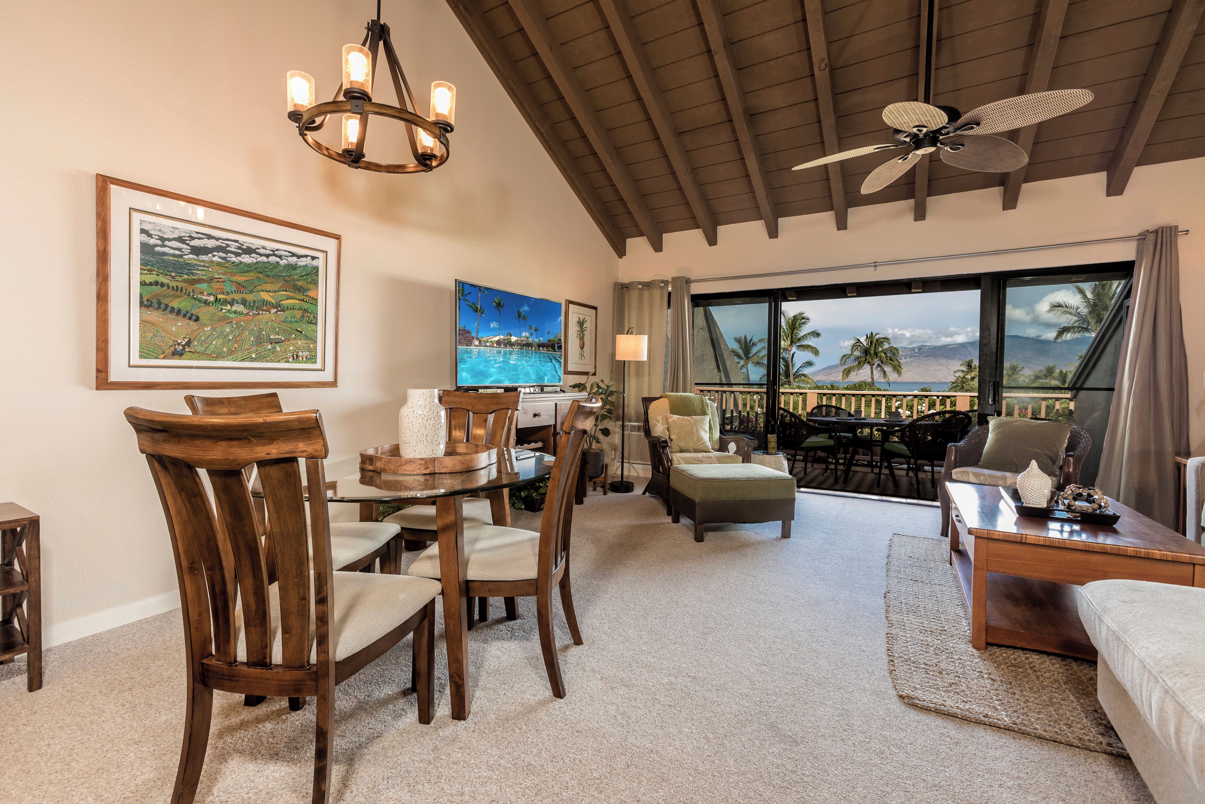A spacious living area with a dining table, chairs, a sofa, and a balcony with a view of palm trees and mountains.